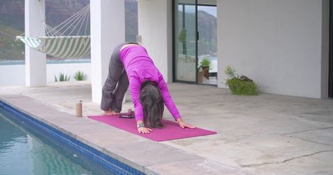 Woman practicing downward dog on pool terrace with hammock and waterfront view