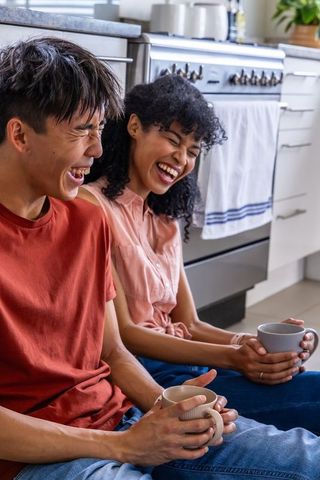 Laughing couple holding mugs on kitchen floor sharing cozy morning coffee, intimate warmth