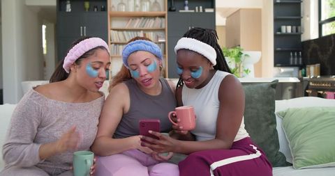 Three friends wearing under-eye gel patches sharing phone and coffee on cozy living room sofa