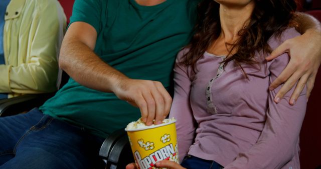 Couple is seated in a movie theater, enjoying a shared bucket of popcorn. Lending a relaxed and intimate atmosphere, this image can be used in contexts such as promoting cinema experiences, relationship articles, entertainment industry, date night ideas, or advertisements for snack foods.