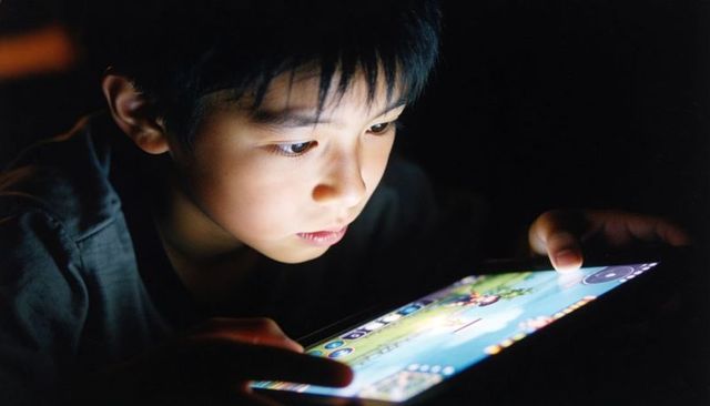 Young Asian boy playing tablet game at night with backlit face and intense concentration