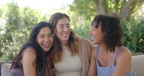 Laughing women sharing moment on sunlit garden sofa, diverse friends enjoying outdoors