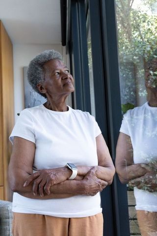 Senior african woman standing by glass door looking up wearing smartwatch reflecting