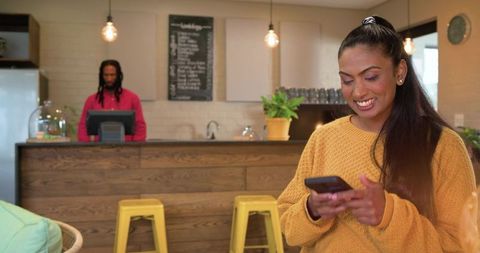 Indian woman smiling while using smartphone in cozy cafe with barista and wooden counter