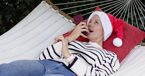 Mature woman relaxing on hammock wearing santa hat and talking on smartphone outdoors