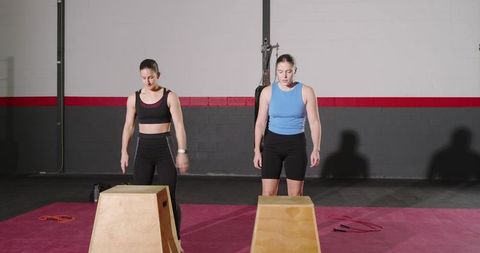 Two women training with plyo boxes in gym performing box jumps and strength conditioning