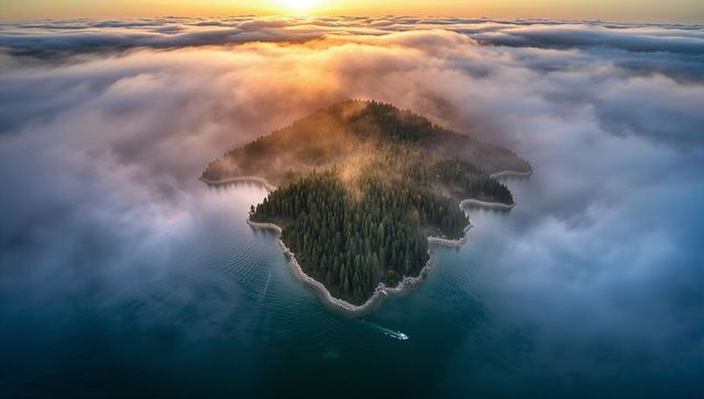 Drone-captured sunrise fog enveloping forested island with rocky shoreline and boat wake