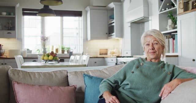 Senior woman is sitting comfortably in a modern kitchen. She is gazing warmly towards the camera, showcasing a tranquil and relaxed atmosphere. This image is great for depicting concepts of elderly life, comfort, and domestic tranquility in contemporary home settings. Ideal for use in promotional material for senior living, health and wellness for the elderly, and home lifestyle magazines.