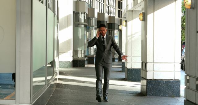 A professional businessman is walking outside an office building, speaking on his smartphone. He is wearing a suit, indicating a formal business environment. This scene can be used for concepts related to corporate life, business communication, or urban work settings. Ideal for illustrating business articles, presentations, and marketing materials.