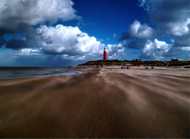 Extensive sandy coastline with an iconic red lighthouse under dramatic cloudscape. Wind patterns visible on the sand create dynamic motion, enhancing the vibrancy of this coastal seascape. Perfect for travel brochures, website banners, or as a scenic background image to evoke a sense of wanderlust, excitement, and coastal serenity.