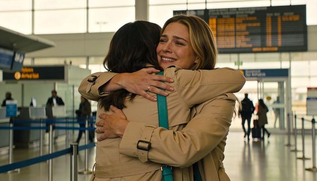 Emotional airport reunion: women hugging in trench coats with teal bag and departure board