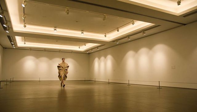 Model walking through minimalist gallery wearing sculptural beige origami coat on runway