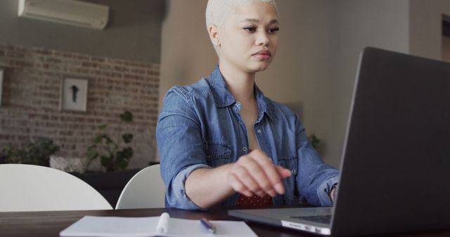 Young professional woman sitting at a desk, working on her laptop in a home office. She looks focused and determined. Suitable for illustrating remote work, productivity, or modern working environments.