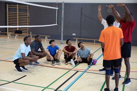 Diverse youth volleyball team listening while coaches demonstrating block technique in gym