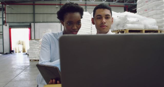 Two employees collaborating over a laptop in a warehouse surrounded by stacked bags and industrial materials. Ideal for showcasing teamwork and collaboration in logistics, inventory management, and industrial settings. Useful for websites or promotions related to warehousing, supply chain, logistics operations, and technology integration in workplaces.