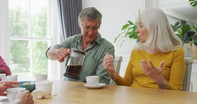 Senior man pouring coffee into cup while enjoying breakfast with elderly woman at bright dining table. Perfect for promoting senior lifestyle products, family gatherings, healthy living, retirement community activities, and home decor services.