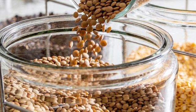 Pouring tan lentils into glass jar on pantry shelf for bulk food storage and display