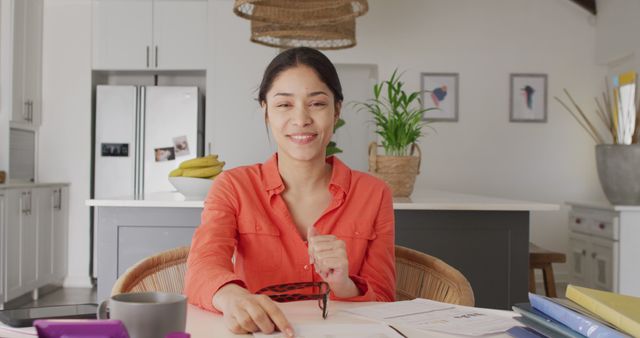 Woman in orange blouse working from a modern kitchen table with documents, laptop, and coffee cup. Ideal for depicting remote work, home office setups, work-life balance, and casual business environments.