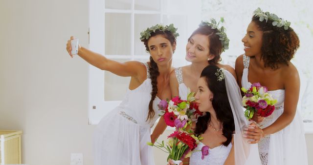Group of bride and bridesmaids wearing white dresses and floral crowns, taking a selfie together. Ideal for wedding-related content, such as wedding planning websites, bridal magazines, and social media posts celebrating wedding moments.