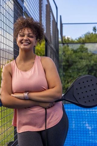 Smiling woman leaning on fence holding padel racket on outdoor court wearing pink top