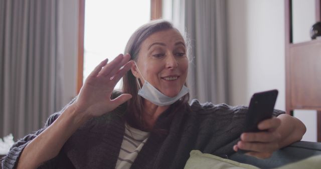 Middle-aged woman sitting on couch, smiling and waving at smartphone while video chatting. She is wearing a mask under her chin, indicating a casual or post-pandemic setting. This can be used for themes related to virtual communication, staying connected with family and friends, or adapting to new norms in personal interactions.