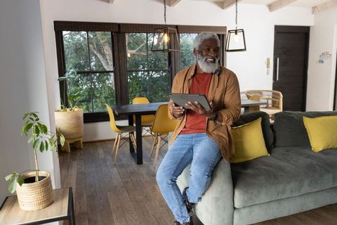 Senior african american man sitting on sofa holding tablet smiling in bright cozy living room