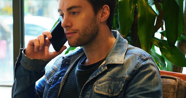 Young man with stubble talking on phone in cafe, wearing denim jacket. Used for illustrating modern communication, young adults in casual settings, urban lifestyle, and business or personal interactions.