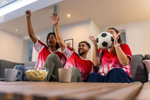 Group of friends cheering while watching soccer match at home with popcorn and ball