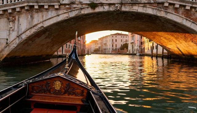 Gondola gliding under stone venetian bridge at sunset with golden water reflections