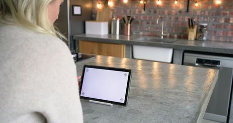 Blonde woman leaning on kitchen countertop tapping tablet with stylus in rustic kitchen