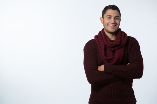 Young man standing with arms crossed, wearing a maroon sweater and matching scarf. He is smiling confidently against a plain white background. Ideal for use in advertisements, lifestyle blogs, fashion promotions, and social media content.