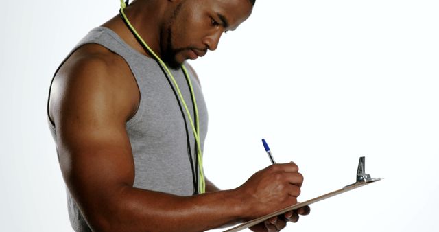 This stock photo depicts a male personal trainer concentrating as he writes a workout plan on a clipboard. He is wearing a grey tank top and has a whistle around his neck. This image is ideal for promoting fitness programs, gym memberships, health and wellness blogs, personal training services, sports advertisements, and fitness-related content.