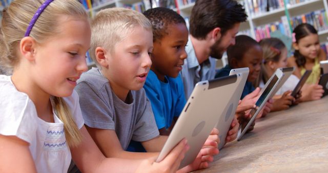 A diverse group of children and a young Caucasian male teacher engage with tablets in a library setting, with copy space. Their focused expressions suggest a learning activity or educational technology use.