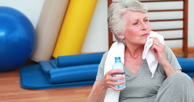 Senior woman in grey workout attire taking a break in a gym setting. She is holding a water bottle and wiping sweat with a towel, indicating activity and dedication to staying fit. The image includes exercise mats and a fitness ball in the background, suggesting a comprehensive workout environment. Ideal for articles on senior health, fitness programs, rehabilitation centers, and lifestyle blogs promoting active living for seniors.