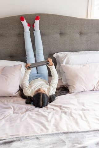 Girl lying upside down on bed with legs up holding tablet, cozy pastel bedroom moment