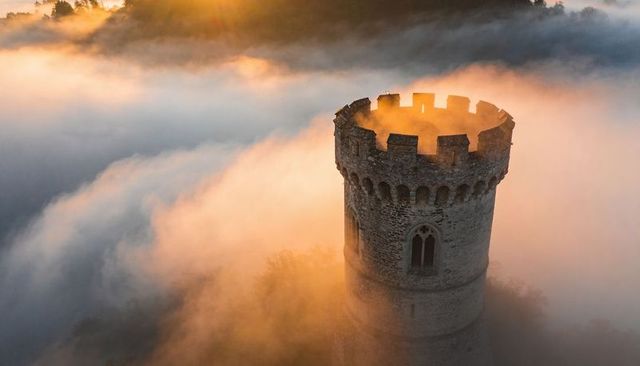 Medieval tower rising through golden morning fog, casting warm light over valley mist