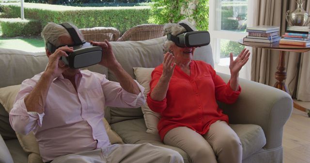 Senior couple sitting on sofa and enjoying virtual reality experience with VR headsets. They appear thrilled and deeply engaged in the immersive world. This image can be used for promoting technology products targeted at elderly users, articles on seniors embracing modern technology, or creators wanting to showcase intergenerational technology adoption. Ideal for blogs, websites, or advertisements centered around senior citizens and tech-savvy lifestyles.