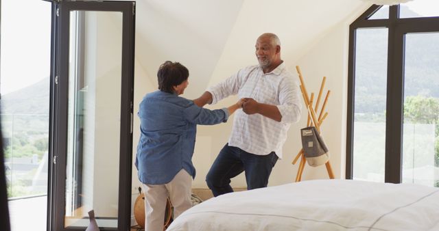 Senior couple dancing together in a spacious, modern bedroom with large windows and a scenic mountain view. Elderly man and woman in casual clothing enjoy a happy moment of leisure and love. This image can be used for portraying active and joyful senior lifestyles, retirement activities, or romantic moments in later years.