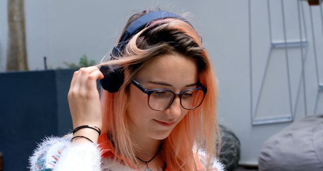 Young woman with pink hair sitting outdoors, listening to music on headphones. She is wearing glasses and casual attire, smiling and enjoying her leisure time. Ideal for websites and advertisements related to lifestyle, youth culture, music streaming services, and tech gadget promotions.