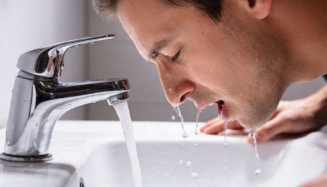 Man rinsing face at bathroom sink with running water from chrome faucet, close-up profile