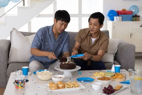 Two asian men celebrating birthday at home cutting chocolate cake on sofa with snacks