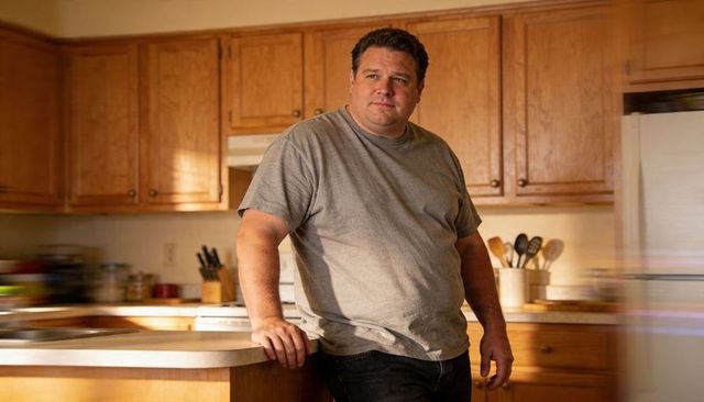 Middle-aged man leaning on kitchen island wearing gray tee, warm cozy home interior