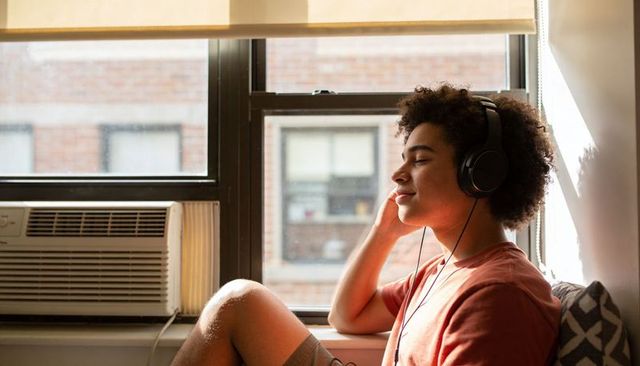 Young man relaxing on sunlit windowsill with headphones in urban apartment