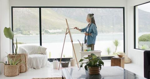 Mature woman painting at easel in bright waterfront living room studio with plants