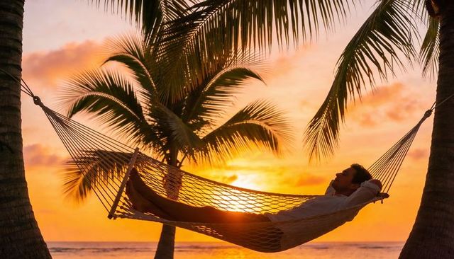 Man relaxing in hammock at tropical sunset between palm trees on tranquil beach