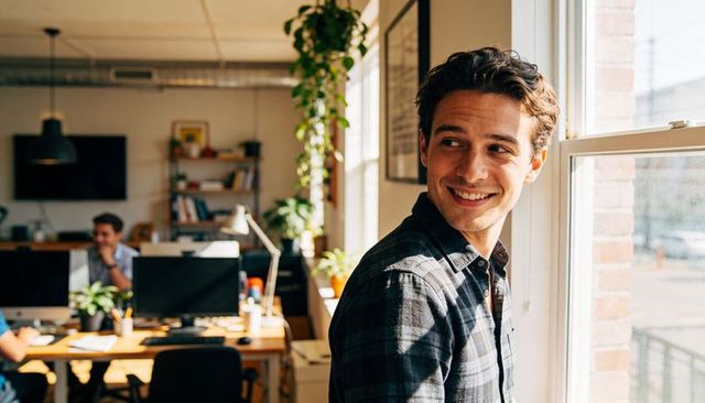 Smiling young professional looking out window in sunlit coworking office, casual plaid