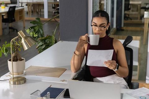 Professional woman sipping coffee while reading document at modern open-plan office desk
