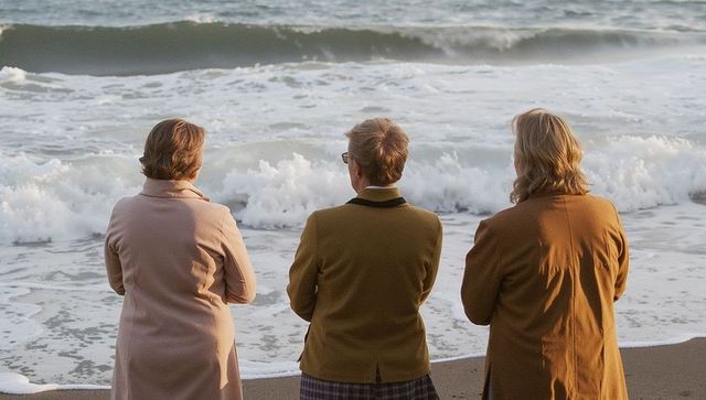 Three women gazing at rough ocean waves on sandy beach wearing autumn coats, contemplative trio