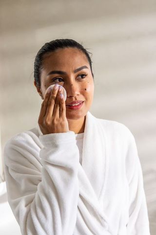 Asian woman removing makeup with cotton pad in plush white bathrobe calm skincare routine