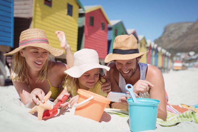 Family lying on sand in front of colorful beach huts, enjoying a sunny day. Parents and child wearing hats, playing with beach toys, and smiling. Perfect for vacation promotions, family bonding themes, summer activities, and travel advertisements.